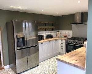 A new modern kitchen installation that features patterned tiled flooring, white wall tiles, white cabinets, wooden countertops, an oven with extractor fan and a large silver fridge.