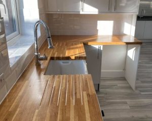 A new kitchen featuring wooden countertops, white tiled walls, white cabinets and cupboards and grey laminate flooring.