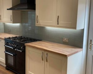 A new kitchen fitted that includes new white cabinets and cupboards, green wall tiles, wooden countertops and a cooker with extractor fan.