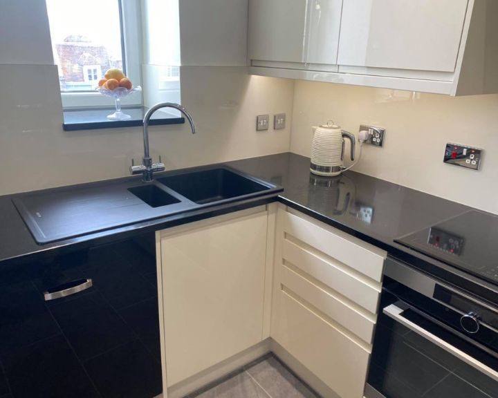 A new residential kitchen installation that features white cabinets and cupboards, a black integrated oven with hob, a black composite sink, black countertops, a grey tiled floor and white wall tiles.