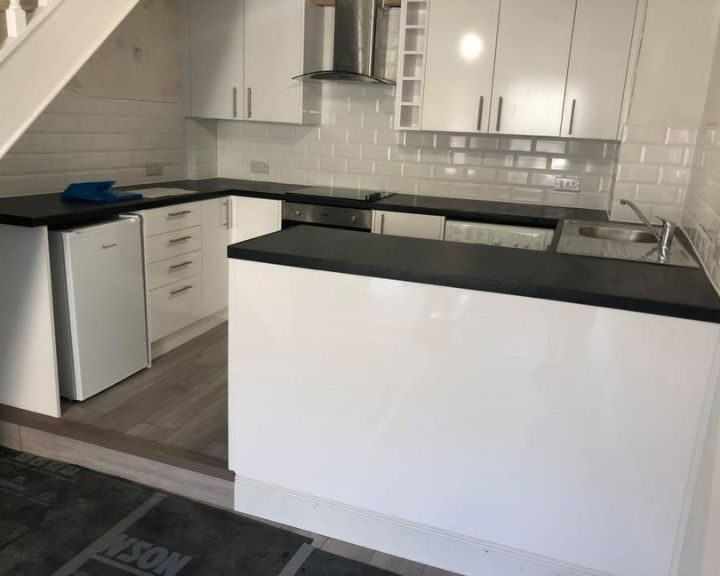 A new kitchen installation that has been fitted under some stairs that features white cupboards and cabinets, white wall tiling, black countertops, a metal sink and integrated cooker with hob and extractor fan.