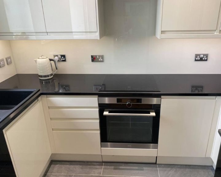 A new residential kitchen installation that features white cabinets and cupboards, a black integrated oven with hob, black countertops, a grey tiled floor and white wall tiles.